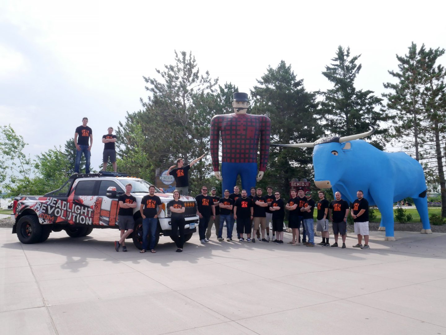 The Headlight Revolution team in matching shirts posing with a branded truck beside the Paul Bunyan and Babe the Blue Ox statues in Bemidji.