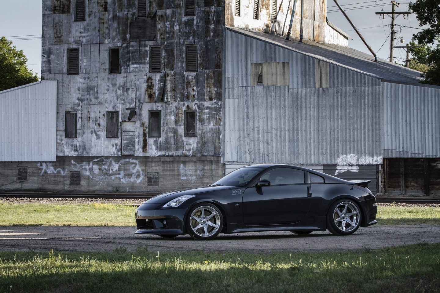 A modified black Nissan 350Z parked in front of a weathered grain elevator.