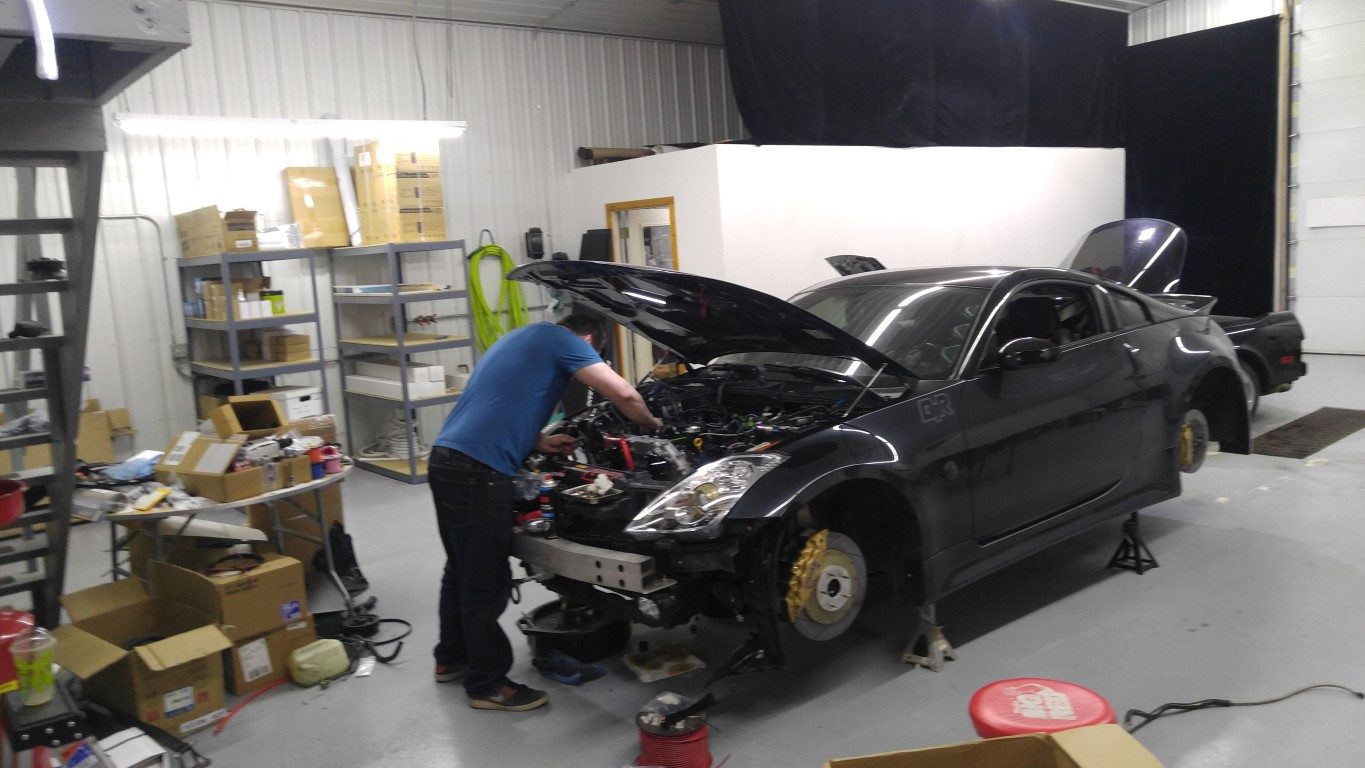 Chris Nelson leaning into the engine bay of his 350Z, front end disassembled, inside a commercial shop in Bemidji.